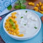 mango raita served in white bowl, garnished with mint and chopped mango. Bowl is kept over a wooden board with mint leaves and cut mango.