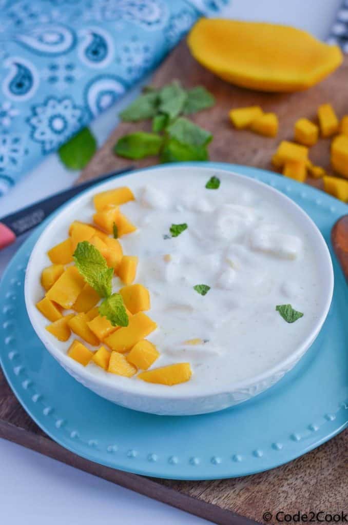 mango raita served in white bowl, garnished with mint and chopped mango. Bowl is kept over a wooden board with mint leaves and cut mango.