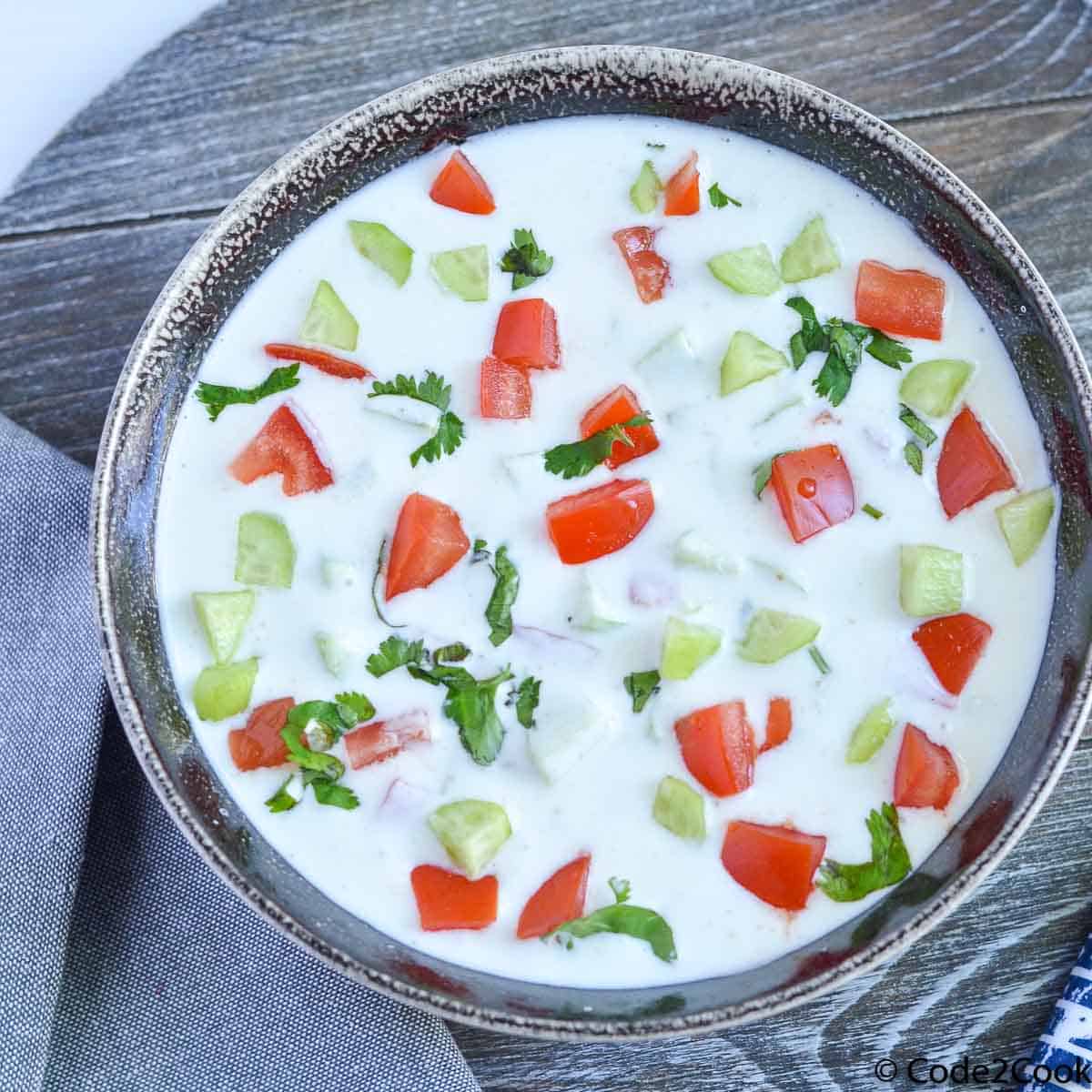 Raita is served in grey bowl and garnished with chopped tomato and cucumber.