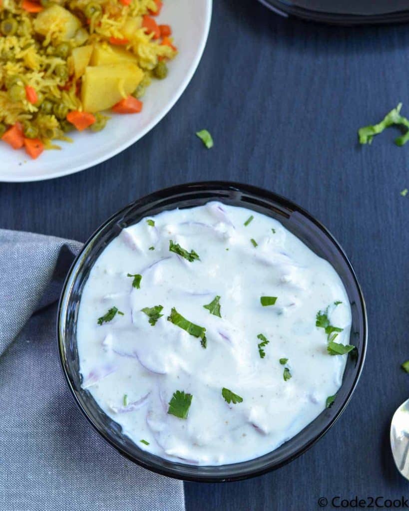 onion raita served in black bowl, garnished with green coriander. Vegetable pulao is served along with the raita.